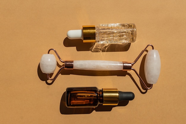 Rouleau de massage pour le visage en quartz rose avec flacon de sérum en or 24 carats et huile essentielle sur fond beige. Le concept de soins de la peau à la maison.