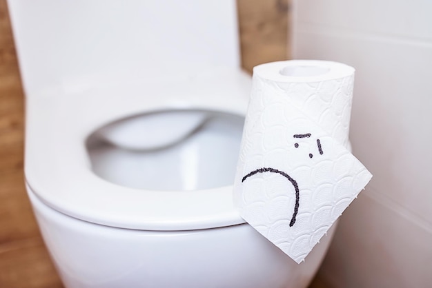 Un rouleau de papier toilette avec un visage triste est suspendu sur les toilettes avec un couvercle ouvert