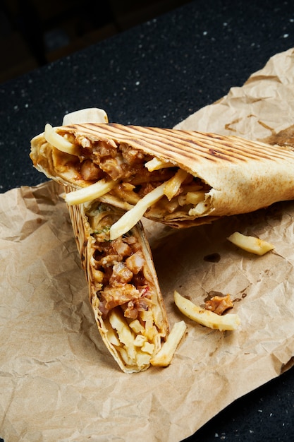 Rouleau de shawarma appétissant avec de la viande, de la salade et de la sauce maison dans un mince pain pita sur du papier kraft sur une surface noire. Cuisine orientale. Kebab en tranches avec de la viande grillée.