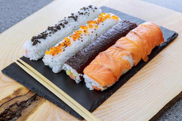 Rouleau De Sushi Au Saumon, Caviar Rouge Et Caviar Noir Sur Une Plaque Noire Sur Une Surface En Bois. Femme à L'aide D'un Tapis Roulant En Bambou Pour Des Sushis Faits Maison