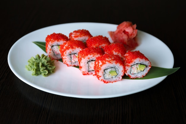 Rouleau de sushi, avec caviar de poisson volant rouge, avocat et fromage à la crème, sur une plaque blanche, sur une surface noire