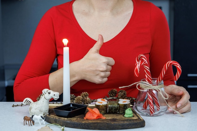 Rouleaux japonais avec du poisson rouge sur une planche à découper en bois Une fille dans un pull rouge mange des sushis avec des baguettes Décoration de table avec des bougies et des bonbons Concept de Noël