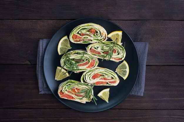 Photo rouleaux de pain pita mince et saumon rouge salé avec des feuilles de laitue sur une plaque en céramique noire, table en bois foncé.