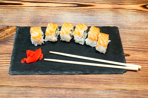 Rouleaux de sushi au poisson avec du saumon, du wasabi et des baguettes sur une planche à découper noire sur un rustique en bois