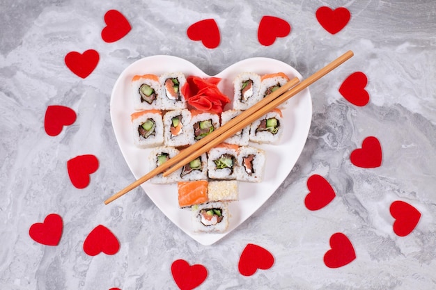 Rouleaux de sushi au saumon sur une assiette blanche en forme de coeur et bâtonnets de sushi en bois parmi les coeurs de confettis rouges