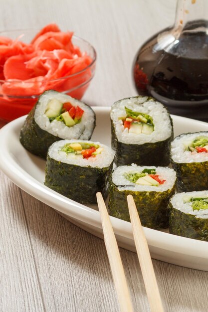 Rouleaux de sushi avec riz, morceaux d'avocat, concombre, poivron rouge et feuilles de laitue sur assiette en céramique, baguettes, bouteille en verre avec sauce soja et gingembre mariné dans un bol. La nourriture végétarienne.