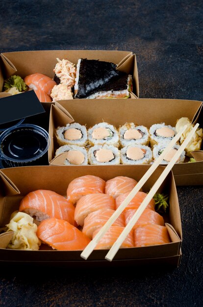 Rouleaux de sushi savoureux dans des boîtes de papier kraft jetables, sauces sur table sombre