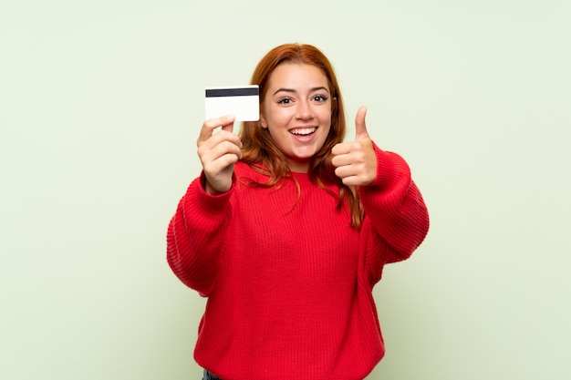 Rousse adolescente avec pull sur vert isolé tenant une carte de crédit