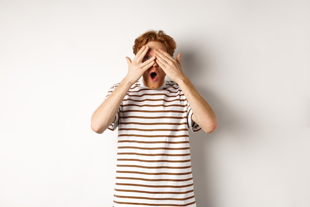 Rousse choqué et embarrassé couvrant les yeux, regardant à travers les doigts et haletant, debout sur fond blanc.