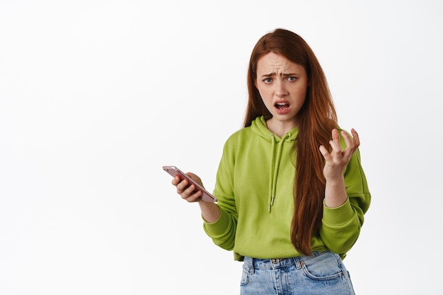 Une rousse frustrée se plaint de quelque chose sur son téléphone portable, a l'air agacée et tient un smartphone, fronçant les sourcils déçu, fond blanc.