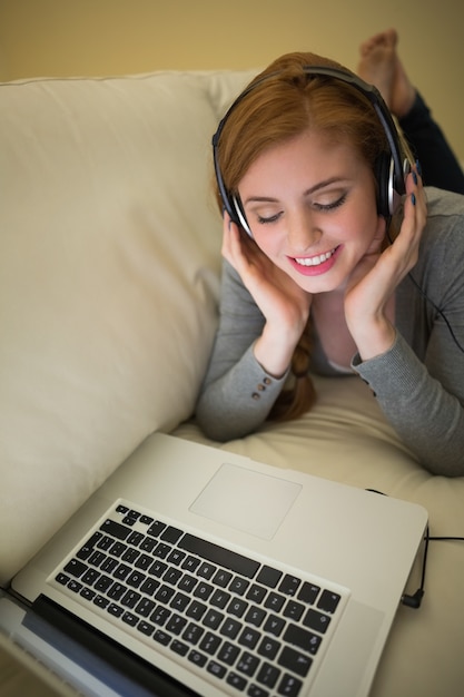 Rousse heureuse allongée sur le canapé avec son ordinateur portable en écoutant de la musique