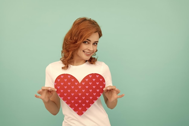 Une rousse joyeuse tient un coeur rouge sur fond bleu