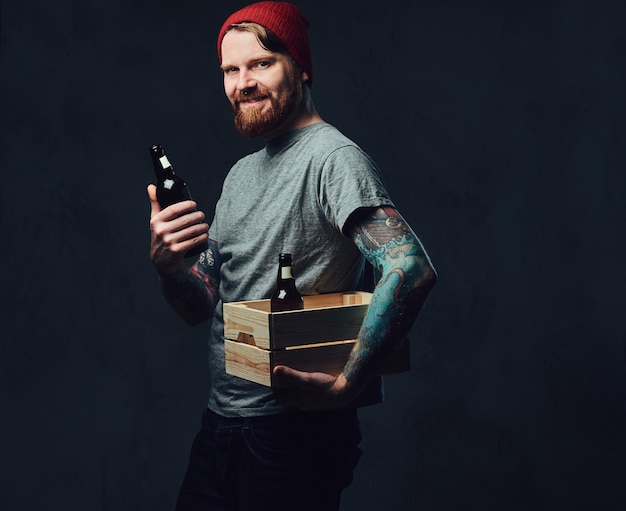 Rousse positive tatouée, un homme barbu tient une boîte en bois avec des bouteilles de bière sur fond gris.