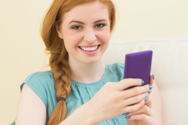 Rousse souriante assise sur le canapé en utilisant un smartphone