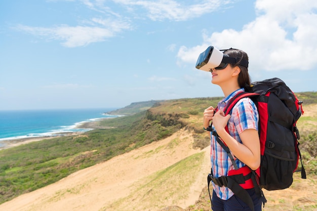 une routarde souriante portant un appareil de réalité virtuelle fait l'expérience d'une vidéo 3D avec un paysage côtier debout sur une montagne de randonnée avec des bagages personnels.