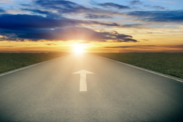La route d'asphalte avec le panneau à flèche blanche va à l'horizon Le soleil à l' horizon et la route s'éloigne en avant Le concept d'avancement Perspectives claires et positives à l'avenir