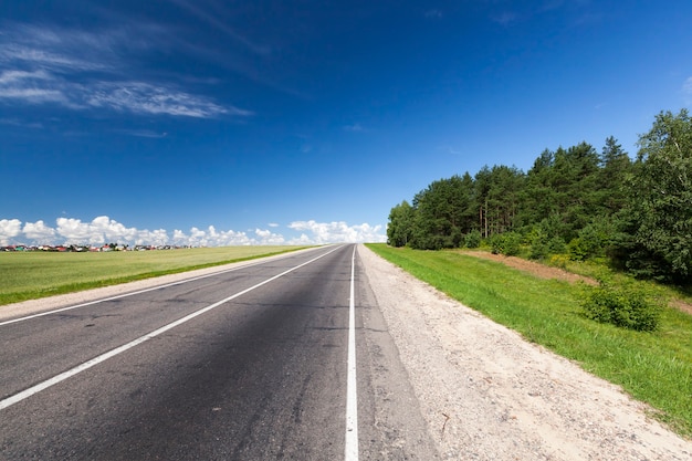 Route asphaltée simple avec bande de marquage blanche pour la régulation de la circulation, paysage d'été avec ciel bleu, herbe et arbres