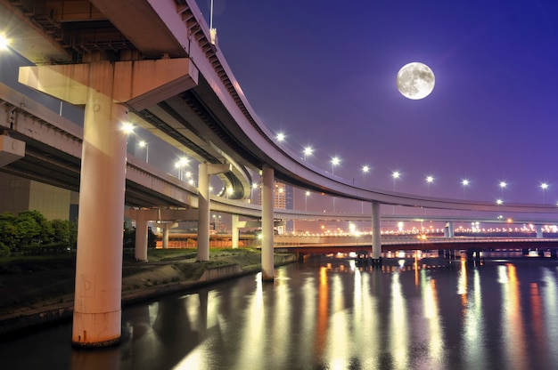 Route d'autoroute suspendue au-dessus des eaux de la baie de Tokyo la nuit avec la pleine lune dans le ciel