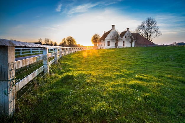 Route de campagne dans la campagne néerlandaise avec une clôture blanche menant vers une vieille ferme en Hollande pendant un coucher de soleil atmosphérique