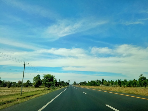 Route de campagne avec rizière jaune, poste électrique, arbre vert et ciel bleu avec des nuages blancs.