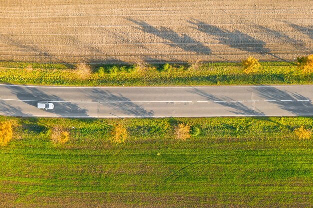 Route entre champ vert et terrain cultivé avec des arbres jaunes au coucher du soleil en automne avec voiture. Vue aérienne sur la piste d'asphalte ou la ruelle des arbres.