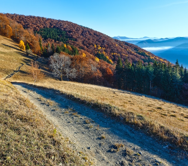 Route rurale en montagne d'automne, forêt colorée sur pente et nuages entre les sommets.