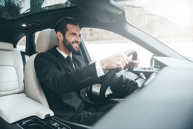 En route vers le succès. Jeune homme d'affaires confiant assis sur le siège avant et souriant en conduisant une voiture