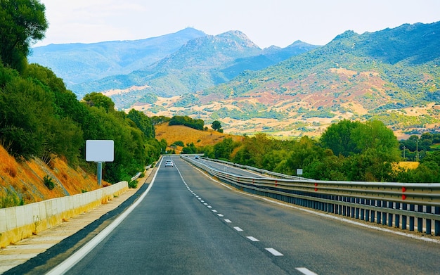 Route vide sans voitures dans l'île de Sardaigne en Italie l'été. Transport roulant sur l'autoroute de l'Europe. Vacances sur autoroute. province de Cagliari. Montagnes en arrière-plan. Technique mixte.