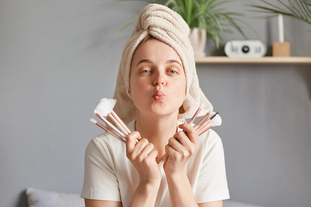 Routine de beauté du matin Jolie jeune femme adulte caucasienne positive portant une molette sur la tête maquillage du matin avec de nouveaux pinceaux cosmétiques envoyant des baisers d'air