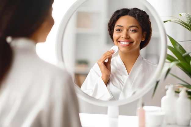 Photo routine de beauté femme noire souriante utilisant un tampon de coton pour nettoyer le visage