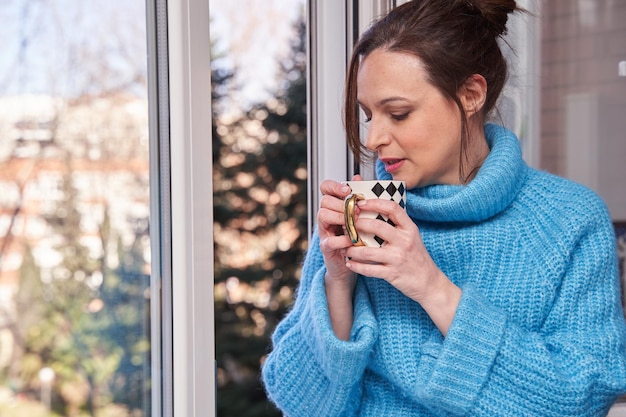 Routine du matin Portrait d'une jeune femme charmante et heureuse, profitant de la vue sur la rue par la fenêtre, buvant un bon café assis sur le rebord de la fenêtre et souriant
