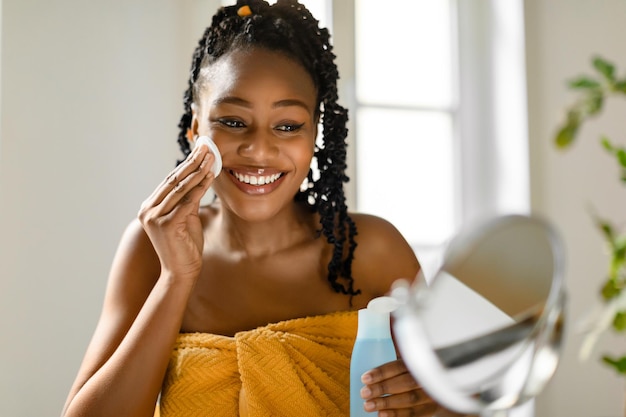 Photo routine de soins du visage heureuse femme noire regardant un miroir à l'aide d'eau micellaire et d'un coton pour le démaquillage