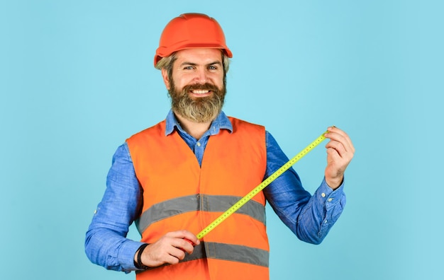 Ruban à mesurer de couleur jaune homme barbu dans un casque travailleur hipster brutal utilise un ruban à mesurer Homme en uniforme travaille sur la production Constructeur de technologie moderne industrielle avec du ruban adhésif