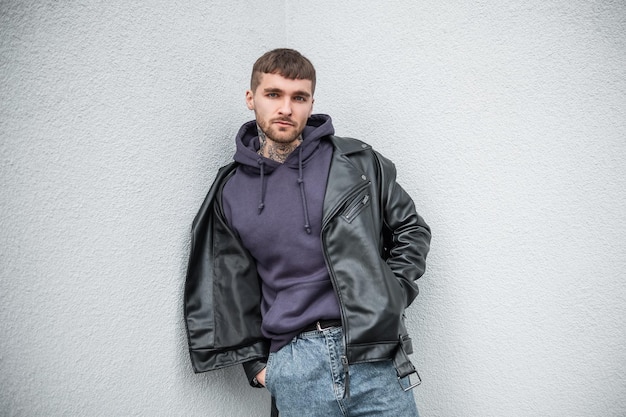 Rue à la mode bel homme hipster dans des vêtements décontractés à la mode avec une veste en cuir et un sweat à capuche se tient près d'un mur blanc dans la rue