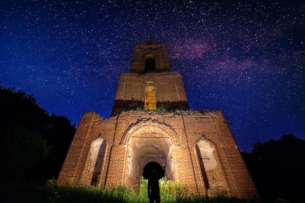 Ruine du clocher de nuit dans la forêt la nuit étoilée et homme avec une lampe de poche en dessous