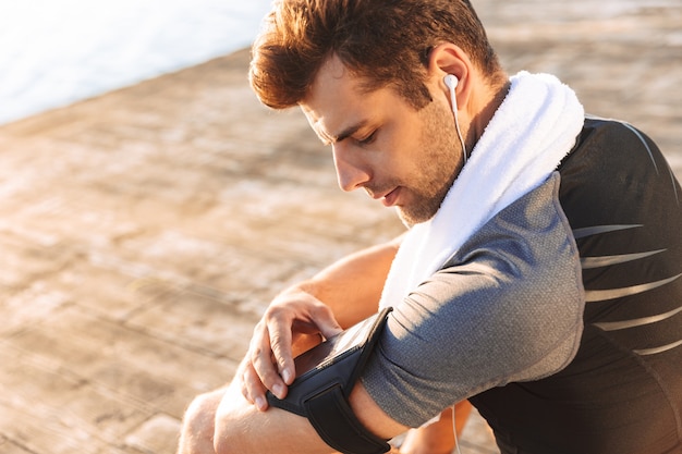 Runner man en survêtement écouter de la musique via un casque sans fil à l'aide d'un support de bras de smartphone tout en étant assis sur une jetée en bois au bord de la mer