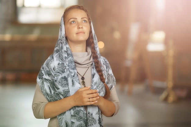 Russe belle femme de race blanche aux cheveux rouges et un foulard sur la tête est dans l'église orthodoxe, allume une bougie et prie devant l'icône.