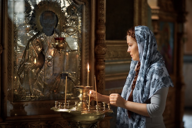 Photo russe belle femme de race blanche aux cheveux rouges et un foulard sur la tête est dans l'église orthodoxe, allume une bougie et prie devant l'icône.