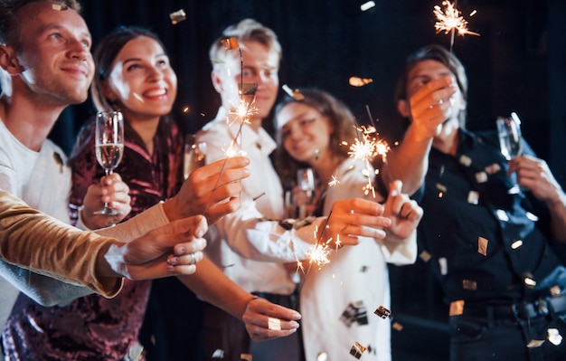 S'amuser avec des cierges magiques. Des confettis sont dans l'air. Groupe d'amis joyeux célébrant le nouvel an à l'intérieur avec des boissons à la main.