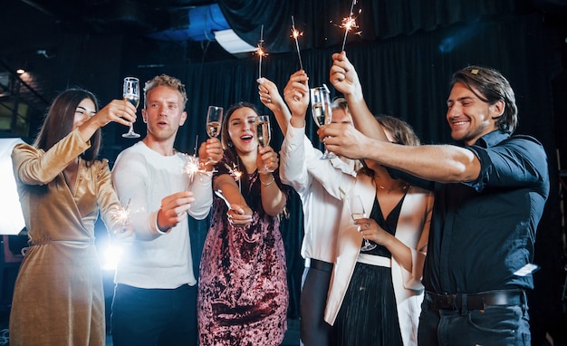 S'amuser avec des cierges magiques. Groupe d'amis joyeux célébrant le nouvel an à l'intérieur avec des boissons à la main.