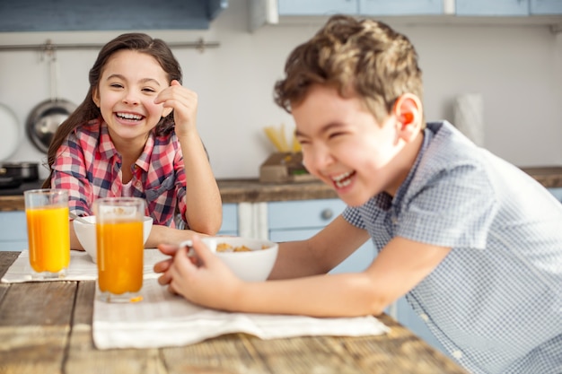 S'amuser ensemble. Jolie petite fille brune riant et prenant son petit déjeuner avec son frère et son frère souriant aussi