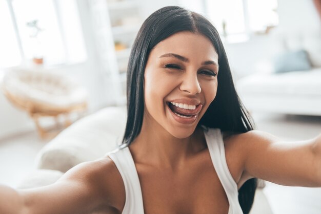 S'amuser à la maison. Autoportrait d'une belle jeune femme regardant la caméra et souriante assise sur le canapé à la maison