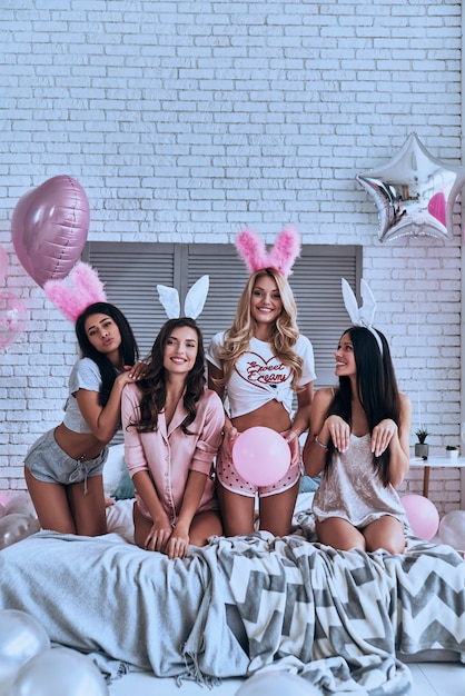 S'amuser. Quatre jeunes femmes souriantes ludiques dans des oreilles de lapin faisant une grimace et faisant des gestes assis sur le lit avec des ballons autour