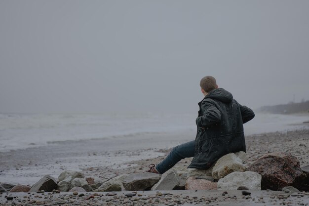 Photo s'assoit dans une veste chaude sur les pierres au bord de la mer sur une plage de sable admire le pittoresque