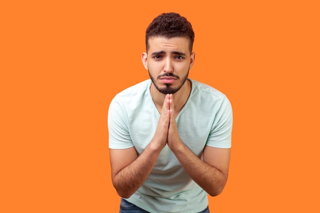 S'il te plaît, je t'en prie. Portrait d'un homme brune bouleversé avec une barbe en t-shirt blanc tenant les bras dans la prière, demandant de l'aide ou pardon avec des yeux implorants. studio d'intérieur tourné isolé sur fond orange