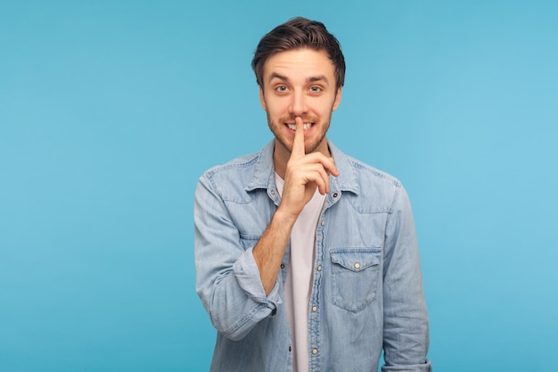 S'il vous plaît, gardez le silence, c'est confidentiel. Portrait d'un homme heureux en chemise denim travailleur montrant un geste de silence avec le doigt sur les lèvres, demandant le secret. studio d'intérieur tourné isolé sur fond bleu