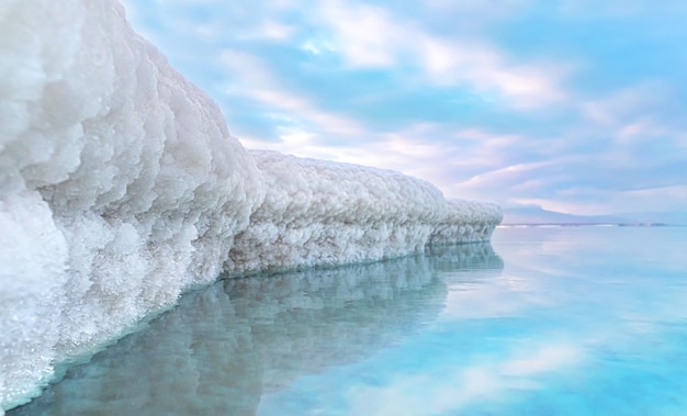 Le sable entièrement recouvert de sel cristallin ressemble à de la glace ou de la neige sur les rives de la mer Morte, de l'eau bleu turquoise à proximité, du ciel coloré avec la distance du soleil du matin - paysage typique de la plage d'Ein Bokek, Israël