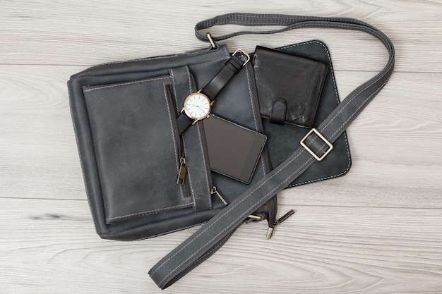 Sac à bandoulière en cuir pour homme avec téléphone portable, montre et portefeuille dessus avec fond en bois gris. Accessoires pour hommes. Vue de dessus.