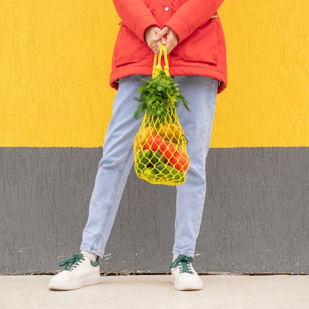 Photo sac à cordes jaune avec concombres, tomates, bananes et herbes dans les mains d'une fille en jean bleu, veste rouge. photo lumineuse dans les tons rouges, jaunes et verts. développement durable, zéro déchet, concept sans plastique.