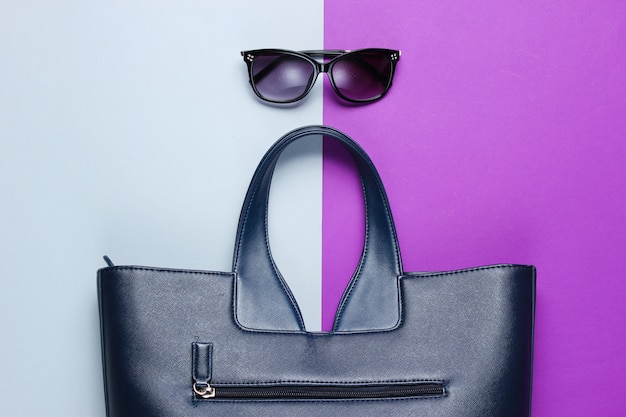 Sac en cuir, lunettes de soleil sur table gris-violet. Vue de dessus